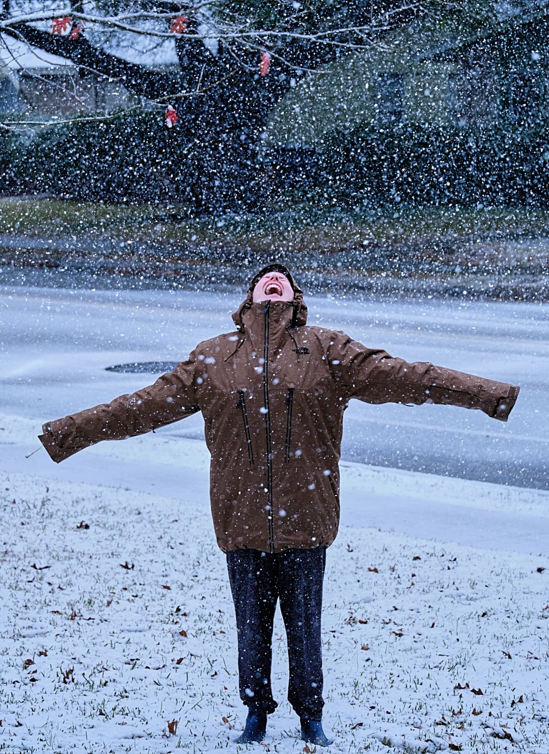 A person in a hooded jacket, arms spread wide, face up to falling snow